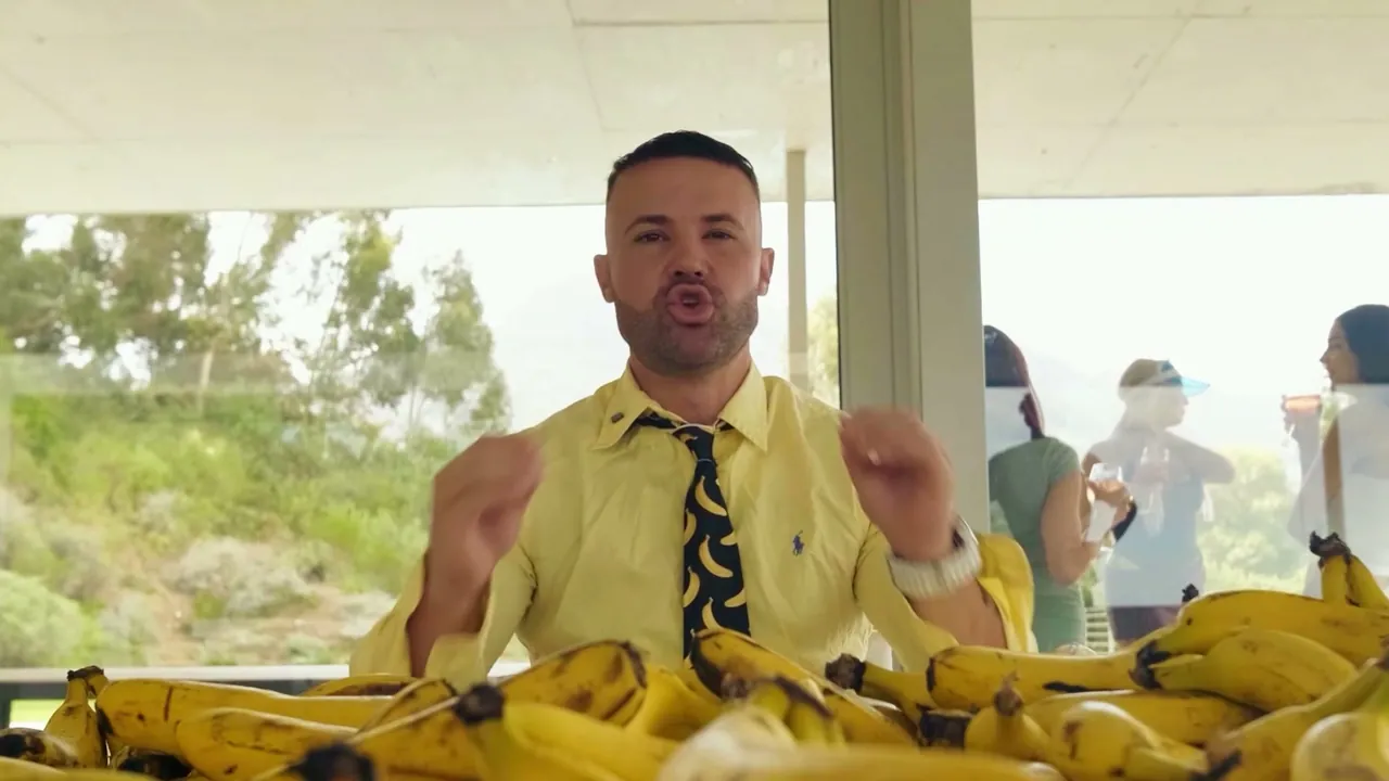 Mike, in a yellow shirt and banana-print tie, talking to camera behind a table piled with bananas.